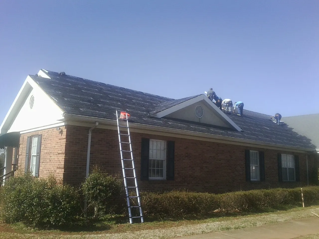 Crew tearing shingles off a brick ranch roof with a ladder set — Emergency Roof Repair work by our crew in Sand Coulee