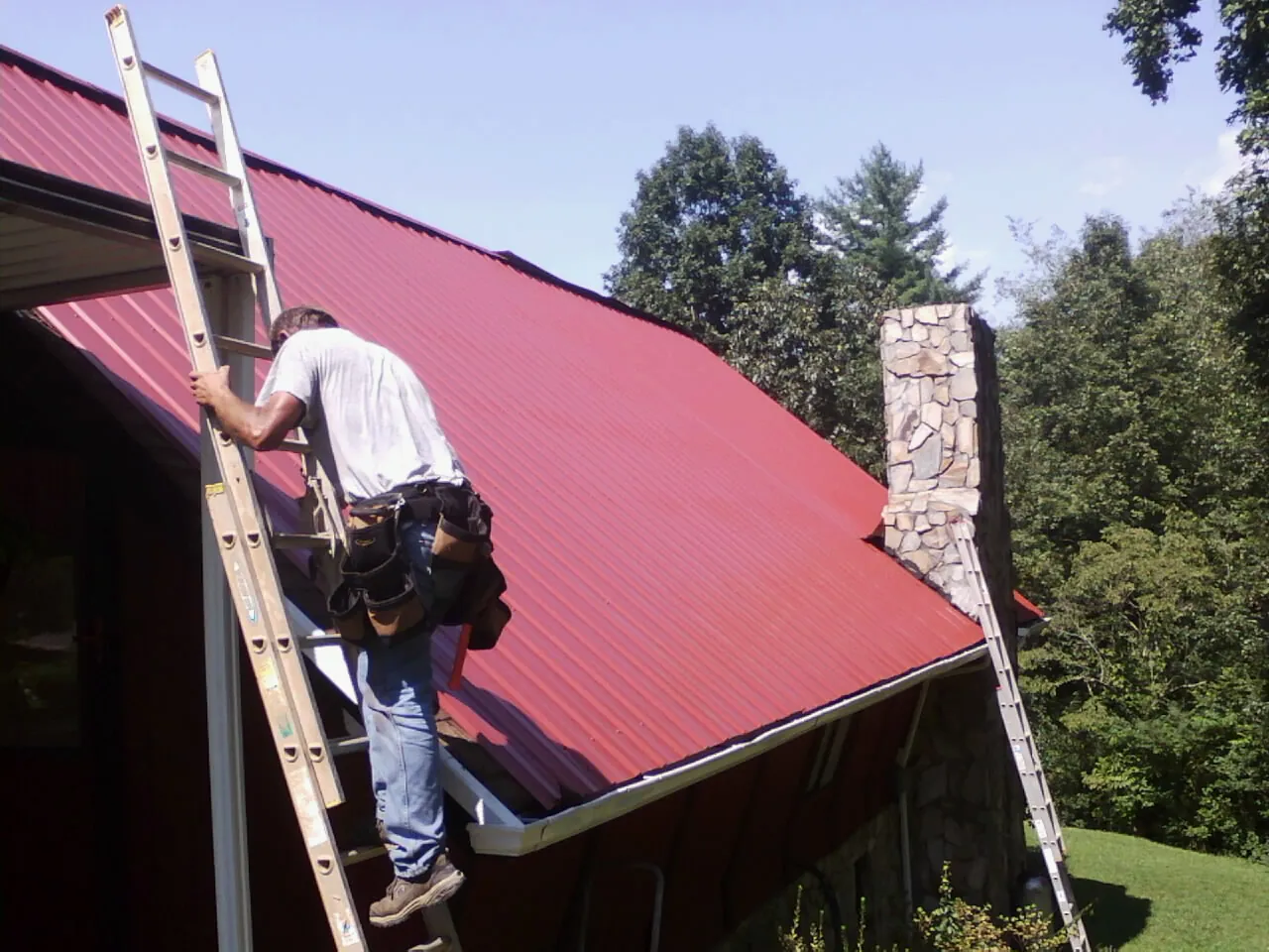 Roofer on a ladder beside a red metal roof and stone chimney — Soffit & Fascia Repair by Roofing Brothers in Sand Coulee